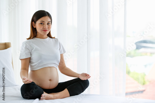 young pretty Asian pregnant woman sitting on a bed doing exercise delightfully by Yoga. She is smiling and looking at a camera. Healthy happy mother concept