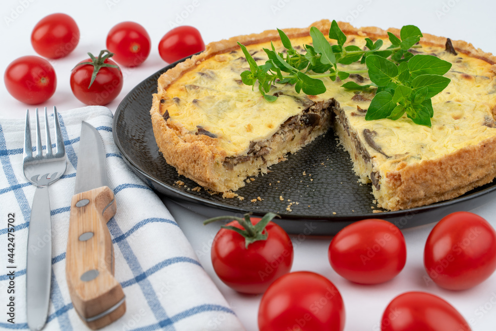 Closeup wide studio shot of freshly baked yellow French salty cake, or quiche with mushrooms, red cherry tomatoes, green mint and kitchen cloth, on black design plate on white background. Cutlery set. Closeup wide studio shot of freshly baked yellow French salty cake, or quiche with mushrooms, red cherry tomatoes, green mint and kitchen cloth, on black design plate on white background. Cutlery set.
