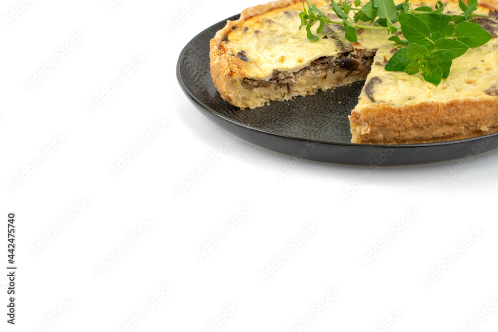 Closeup wide studio shot of freshly baked yellow French salty cake, or quiche, with mushrooms and green mint, on a black design plate, isolated on white background. One slice removed.