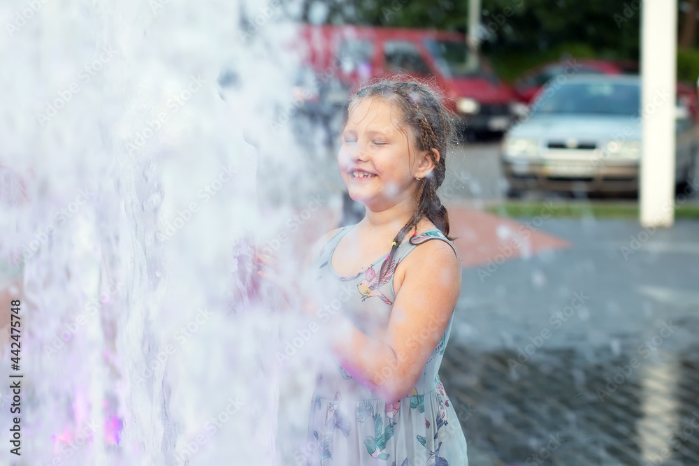 Obraz premium Little cute funny girl child bathes on a hot summer day in a public city fountain. The child runs, plays among the jets and splashes of water