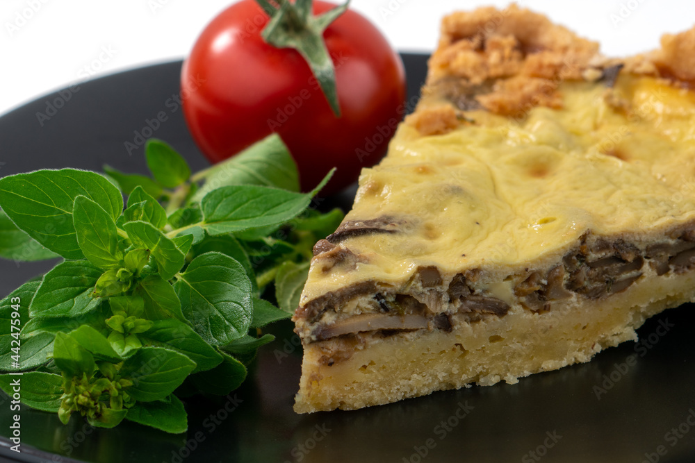Closeup wide studio shot of a slice of yellow French salty cake, or quiche, with mushrooms on a black design plate, isolated on white background. Cherry tomatoes, green mint