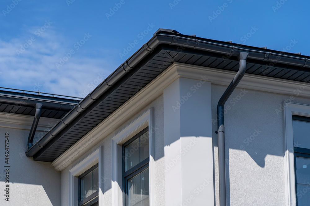 Corner of house with windows, new gray metal tile roof and rain gutter. Metallic Guttering System, Guttering and Drainage Pipe Exterior Corner of house with windows, new gray metal tile roof and rain gutter. Metallic Guttering System, Guttering and Drainage Pipe Exterior