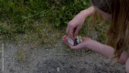 A blonde girl picks up seashells at the side of the road. Grass, asphalt and a young girl squatting. Hand full of seashells.