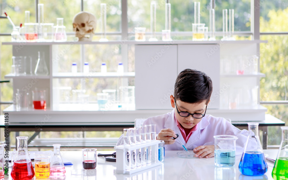 Focused smart Asian schoolboy in lab coat and glasses examining samples while studying chemistry in modern laboratory
