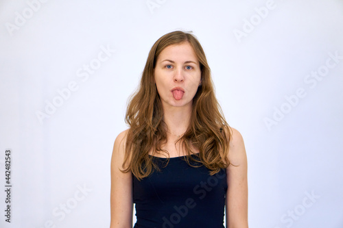 The woman shows her tongue. beautiful Caucasian woman stands on a white background in a blue sweater with loose hair. The woman shows emotions.