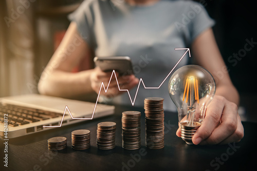 Business women hand holding lightbulb and smartphone with coins stack on desk. concept saving energy and money at office.