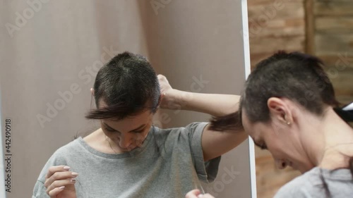 Woman stands in front of a mirror and shave hair bald with the back of head with an electric machine for a haircut.