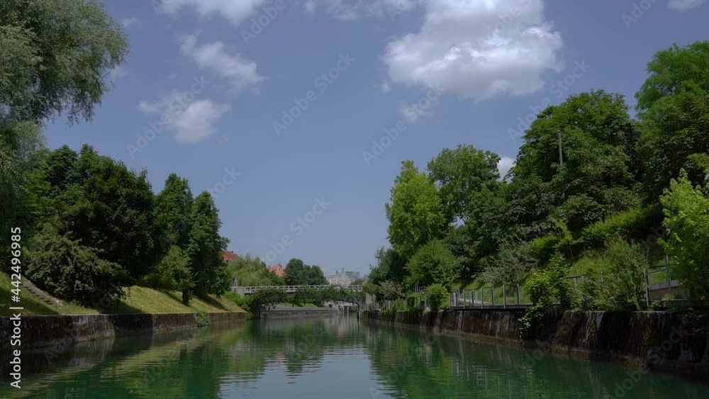 View of Ljubljana city from the boat, Navigating on Ljubljanica river. Bridge and buildings in the distance. Green river bank. Capital of Slovenia in hot summer day. Slow motion, forward moving View of Ljubljana city from the boat, Navigating on Ljubljanica river. Bridge and buildings in the distance. Green river bank. Capital of Slovenia in hot summer day. Slow motion, forward moving
