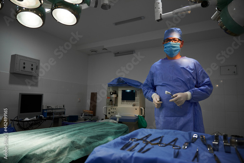 Portrait of plastic surgeon holding medical instruments, standing in operating room. Man doctor wearing surgical uniform, sterile gloves, protective face mask and medical cap. Concept of medicine