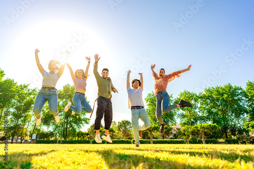 Backlit photography of multiracial group of college students jumping high with open up arms smiling looking at camera in a sunny day with blue sky at city park. Concept of better success in teamwork