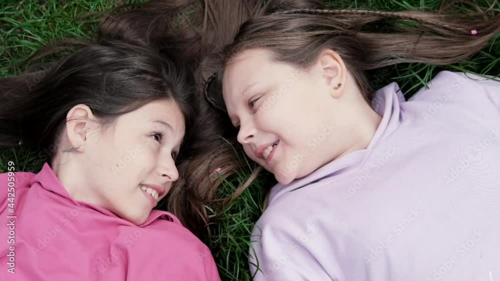 Two pretty millennial kids friends lying on green grass in park or lawn. Caucasian girls smiling and having fun together. Footage from the top view. Two pretty millennial kids friends lying on green grass in park or lawn. Caucasian girls smiling and having fun together. Footage from the top view.
