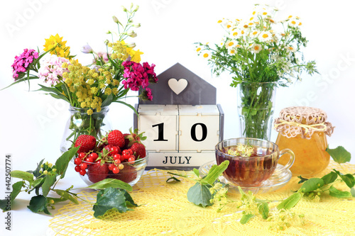 Calendar for July 10 : the name of the month of July in English, cubes with the number 10, bouquets of wild flowers, jam, fruit, a cup of tea on a yellow openwork napkin © MARYIA