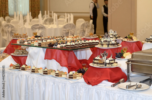 Wallpaper Mural Catering table with a red and white tablecloth with a lot of sweet snacks, small cakes with cream and chocolate cream in a large banquet hall in the background and blurred waiters Torontodigital.ca