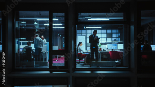 Shot from Outside the Window: Businessmen and Businesswomen Working in the Office. Managers Doing Financial Business in the Evening. Employees Work on Computers, Laptops, Mobile Phones, Tablets.