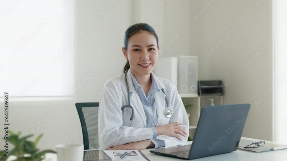 Young adult asia people or medic staff woman sitting toothy smile arm cross look at camera happy work on desk in clinic office room in telehealth telemedicine smart health care consult online service. Young adult asia people or medic staff woman sitting toothy smile arm cross look at camera happy work on desk in clinic office room in telehealth telemedicine smart health care consult online service.