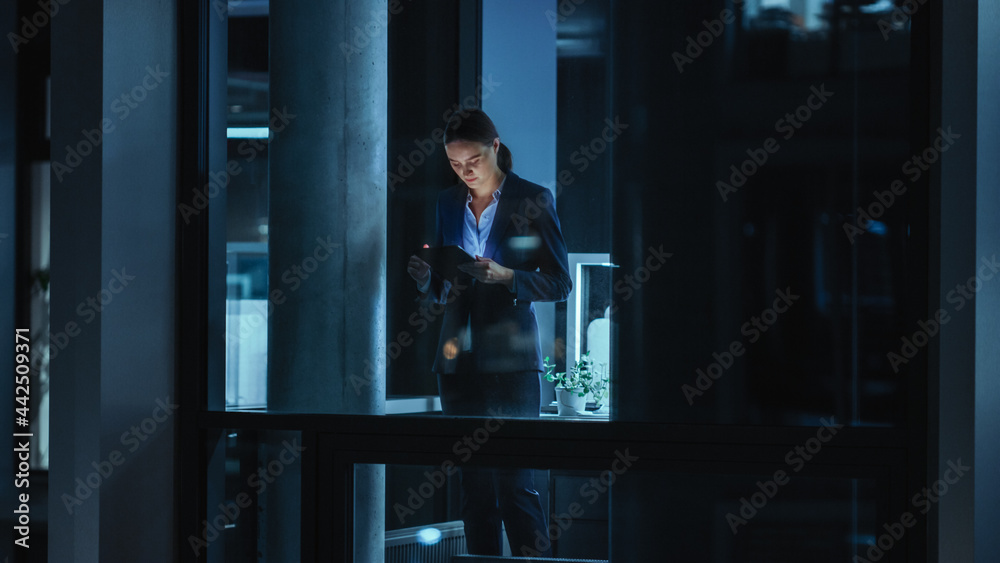 Shot from Outside: Businesswoman Working on a Tablet Computer in the Office. Manager Checking Emails while Standing by the Window. Employee Planning Financial Tasks and Business Development. Shot from Outside: Businesswoman Working on a Tablet Computer in the Office. Manager Checking Emails while Standing by the Window. Employee Planning Financial Tasks and Business Development.