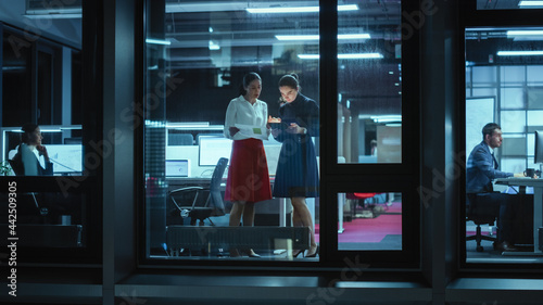 Shot from Outside: Two Businesswomen Working on a Tablet Computer in Office. Managers Standing by the Window. Employees Talk about Financial and Business Development. Colleagues Work Behind Them.