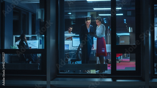 Shot from Outside: Two Businesswomen Working on a Tablet Computer in Office. Managers Standing by the Window. Employees Talk about Financial and Business Development. Colleagues Work Behind Them.