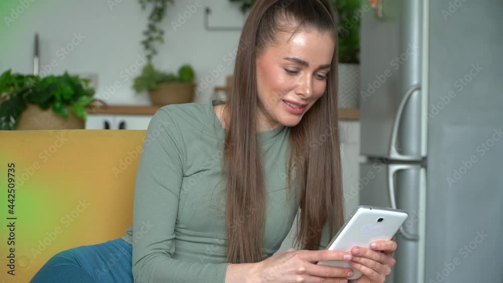 Young smiling caucasian woman sits on sofa at home and uses digital tablet, types message. Online chat. Surfing internet Young smiling caucasian woman sits on sofa at home and uses digital tablet, types message. Online chat. Surfing internet
