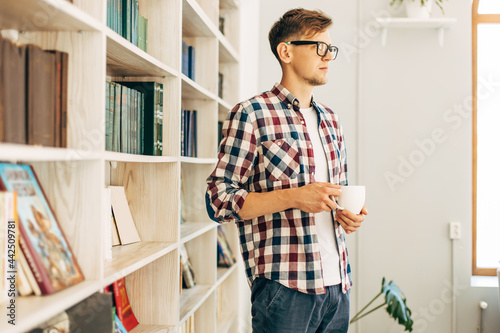 student in glasses and a plaid shirt drinks coffee and relaxes sitting at the table against the background of the wall with books