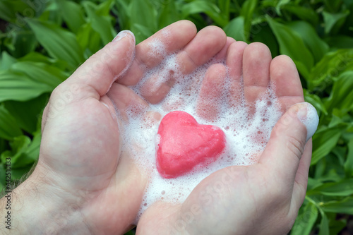 Men's hands hold pink soap with foam in the shape of a heart on a background of greenery. Men's pure love.