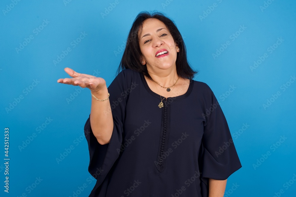 middle aged Arab woman standing against blue background smiling friendly offering something with open hand or handshake as greeting and welcoming. Successful business. middle aged Arab woman standing against blue background smiling friendly offering something with open hand or handshake as greeting and welcoming. Successful business.