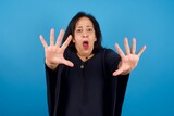 Dissatisfied middle aged Arab woman standing against blue background frowns face, has disgusting expression, shows tongue, expresses non compliance, irritated with somebody.