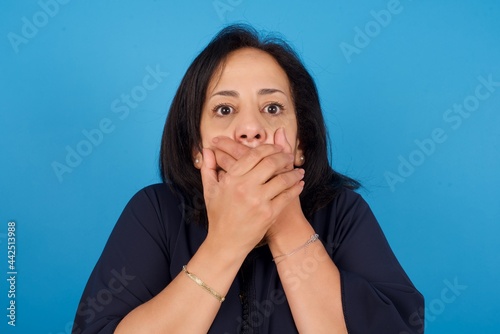 Stunned middle aged Arab woman standing against blue background covers mouth with both hands being afraid from something or after hearing stunning gossips.