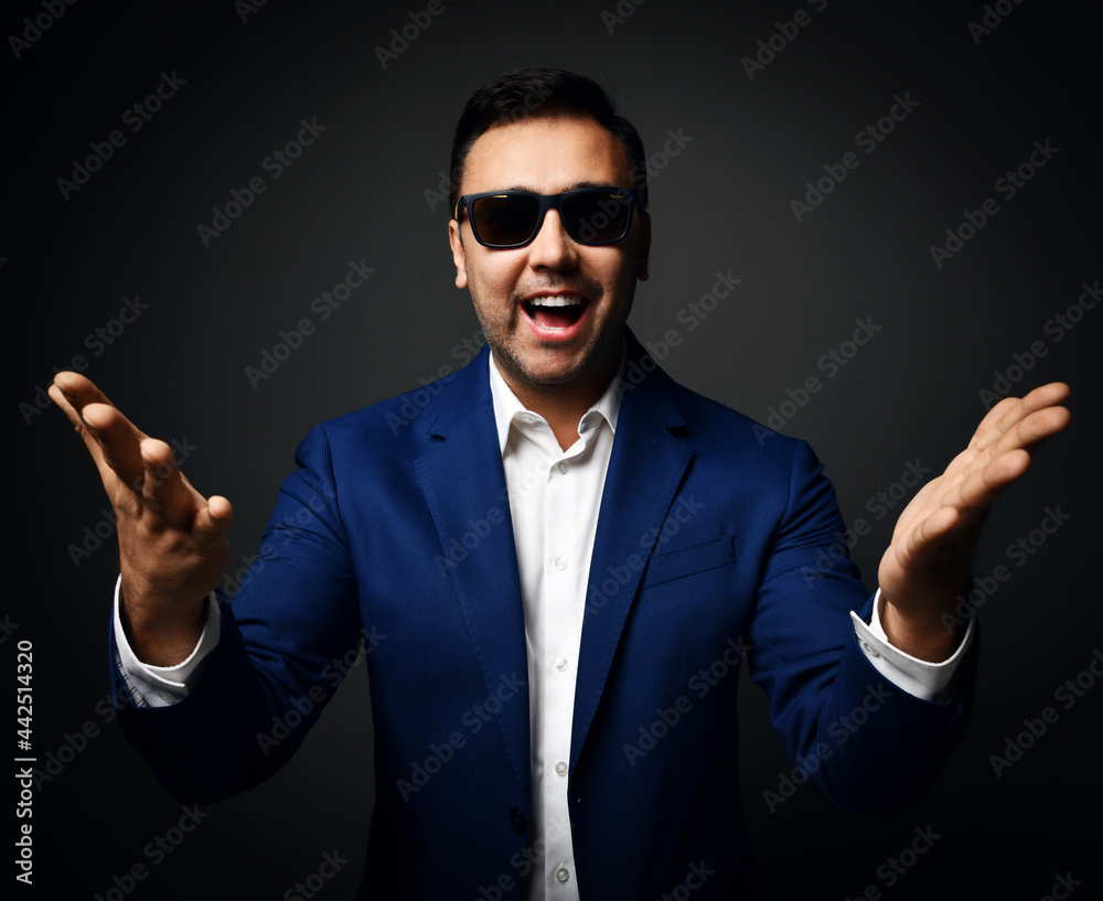 Happy laughing brunet man in blue jacket, white shirt and sunglasses stands holding hands with open palms up at camera over dark background Happy laughing brunet man in blue jacket, white shirt and sunglasses stands holding hands with open palms up at camera over dark background