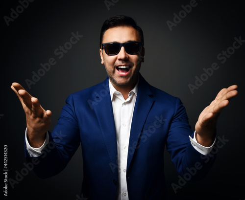 Happy laughing brunet man in blue jacket, white shirt and sunglasses stands holding hands with open palms up at camera over dark background