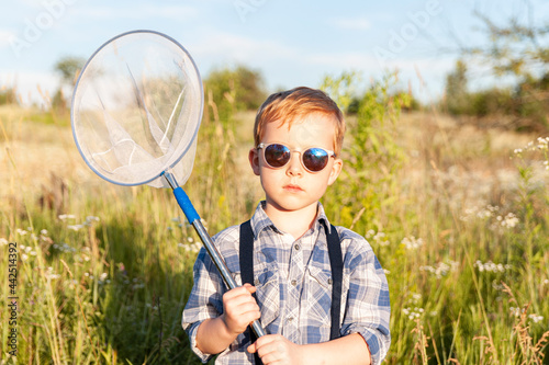 Butterfly hunter - little cute boy wearing sunglass and holding bug net in the hands, standing in the meadow