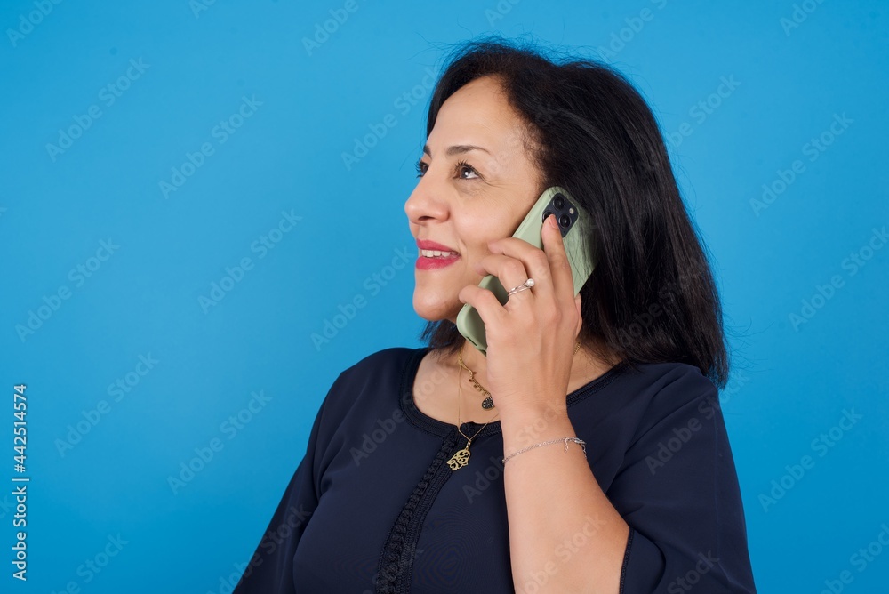 Pleasant looking happy middle aged Arab woman standing against blue background has nice telephone conversation and looks aside, has nice mood and smiles positively while talks via cell phone Pleasant looking happy middle aged Arab woman standing against blue background has nice telephone conversation and looks aside, has nice mood and smiles positively while talks via cell phone