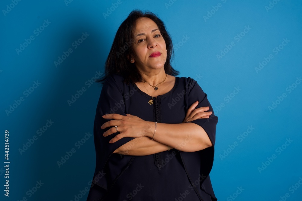 Waist up shot of self confident middle aged Arab woman standing against blue background has broad smile, crosses arms, happy to meet with colleagues. Waist up shot of self confident middle aged Arab woman standing against blue background has broad smile, crosses arms, happy to meet with colleagues.