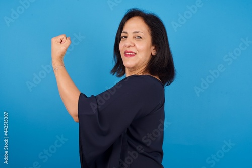 middle aged Arab woman standing against blue background,  showing muscles after workout. Health and strength concept. photo