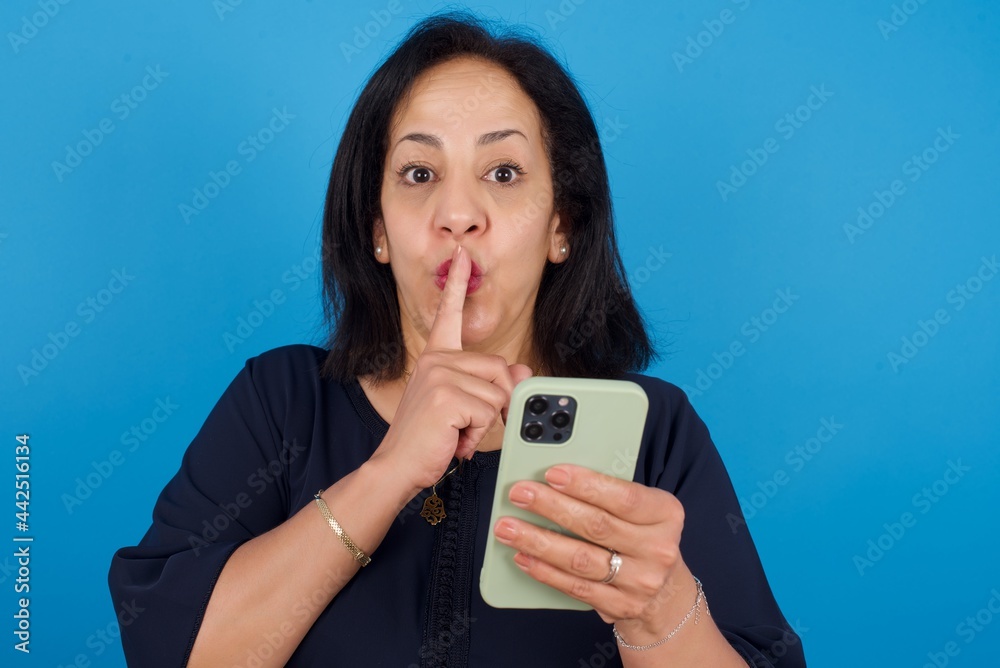 middle aged Arab woman standing against blue background holding modern gadget ask not tell secrets middle aged Arab woman standing against blue background holding modern gadget ask not tell secrets