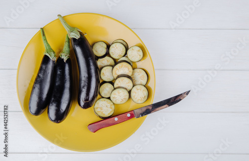 top view of  fresh eggplant with chopped slices and kitchen knife on a yellow plate on white rustic background