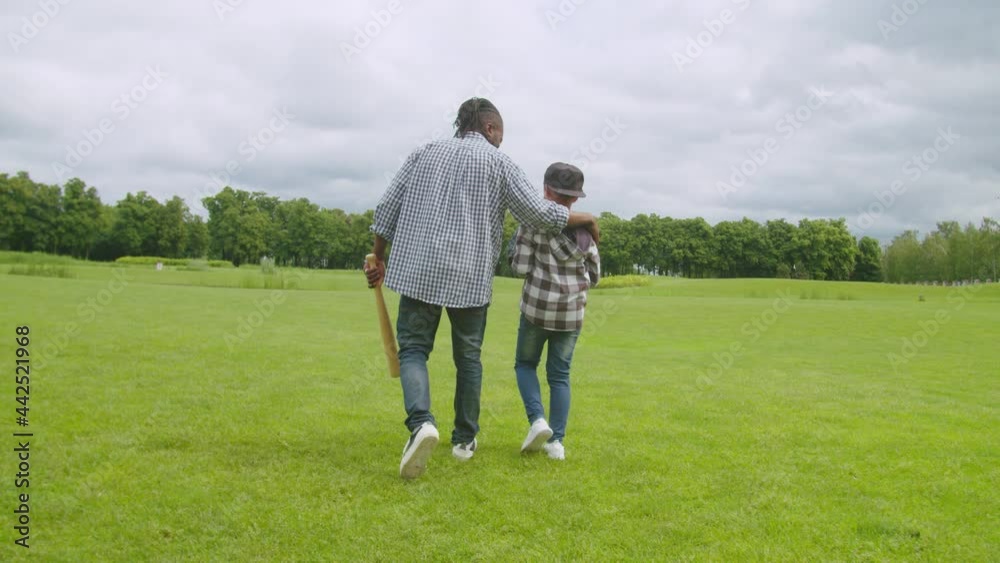 Rear view of attractive african father with dreadlocks and school age boy with baseball glove embracing, friendly chatting and enjoying active lifestyle while walking on green field before baseball. Rear view of attractive african father with dreadlocks and school age boy with baseball glove embracing, friendly chatting and enjoying active lifestyle while walking on green field before baseball.
