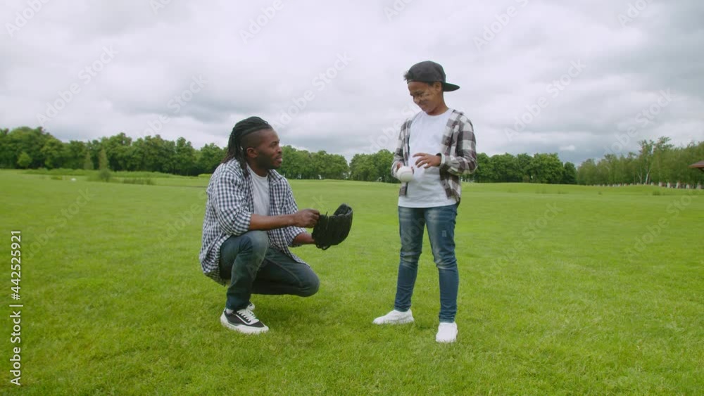 Caring affectionate handsome african american dad with baseball glove sitting in squat position, talking and sharing with cute preadolescent son about baseball while family enjoying leisure in nature.