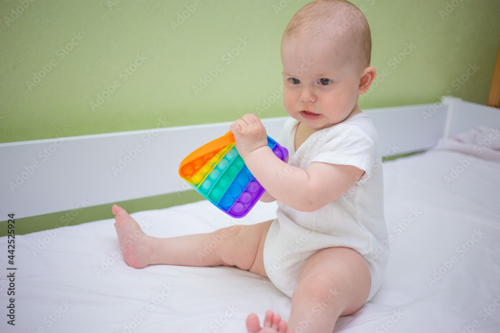 Little baby girl sitting at home on the bed, playing Pop-it new anti-stress toy, popular with children. Kid development idea. Little baby girl sitting at home on the bed, playing Pop-it new anti-stress toy, popular with children. Kid development idea.