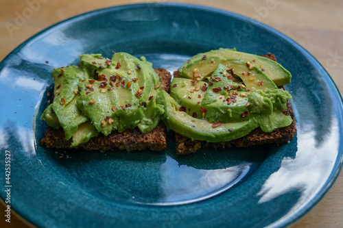 Sliced avocado on brown bread on dark turquoise ceramic plate, chili flakes sprinkled on top.