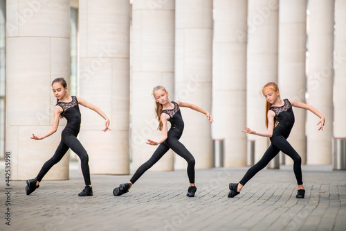 group of three little ballerinas in black tight-fitting costumes are dancing against background of cityscape. © Maria Moroz