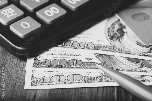 Dollars in one-hundred-dollar bills are spread out on the table next to a calculator, a credit card, and a pen. One-hundred-dollar bills. US dollars. Black and white style.