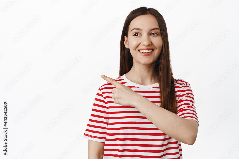 Waist up portrait of brunette 20s girl smiling happy, pointing and looking left at sale, showing banner promo discounts aside, empty space, standing against white background Waist up portrait of brunette 20s girl smiling happy, pointing and looking left at sale, showing banner promo discounts aside, empty space, standing against white background