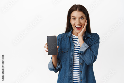 Surprised and happy woman showing smartphone empty screen, phone inferface and look amazed, touching face in disbelief, smiling cheerful, white background photo