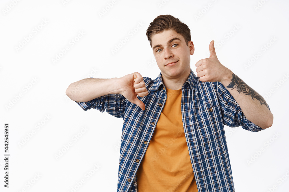 Feedback and rating concept. Young man shows like dislike gesture, thumbs up and down, dont know, average medium quality, standing against white background Feedback and rating concept. Young man shows like dislike gesture, thumbs up and down, dont know, average medium quality, standing against white background