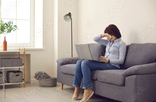 Stressed unhappy woman feeling exhausted after laptop computer overwork at home. Tired overwhelmed suffering from dry eyes syndrome covering face with hand sitting on sofa. Freelance stress, fatigue