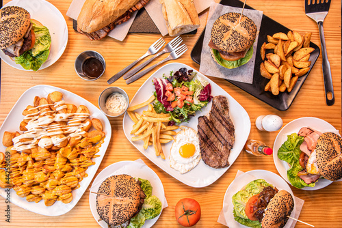 Set of popular Spanish dishes and hamburgers on wooden table. Loin and cheese sandwich, steak and egg, patatas bravas, fresh tomatoes, forks, deluxe potatoes