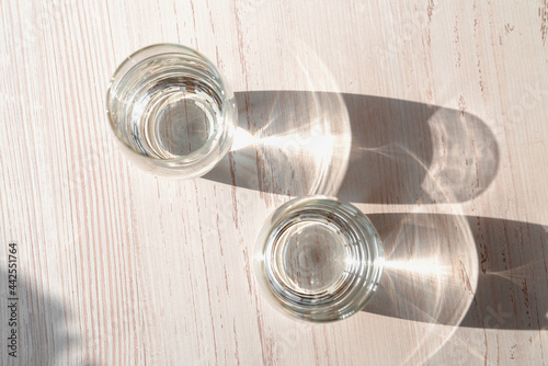 Two glasses of water on white wooden background under direct sun shines. Top view of glasses water. Caustic reflection effect.