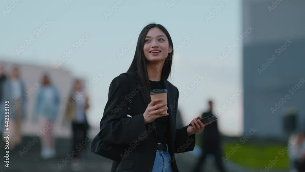 Happy brunette mixed-race young asian woman with coffee walking in the city, using smartphone and drinking hot takeaway coffee. Ethnicity. Urban lifestyle.