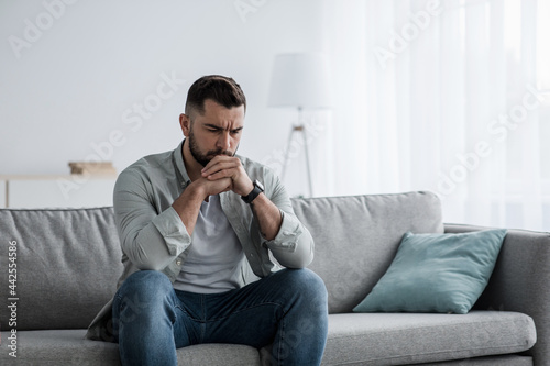 Thoughtful young serious handsome man sit on sofa at home, lost in thoughts, thinking about problem solving